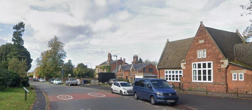 A red 20mph limit painted on a road surface outside a village hall, with cars parked at the side of the road