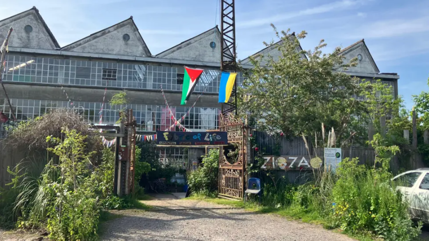 A former factory building with an angular shaped roof and overgrown greenery. The flag of Ukraine and Palestine are flown at the front of the building