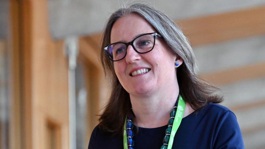 Maree Todd walking down a corridor in the Scottish Parliament. She has shoulder-length hair and glasses, and is wearing a blue top, a blue and green necklace and a lanyard with a green strap.