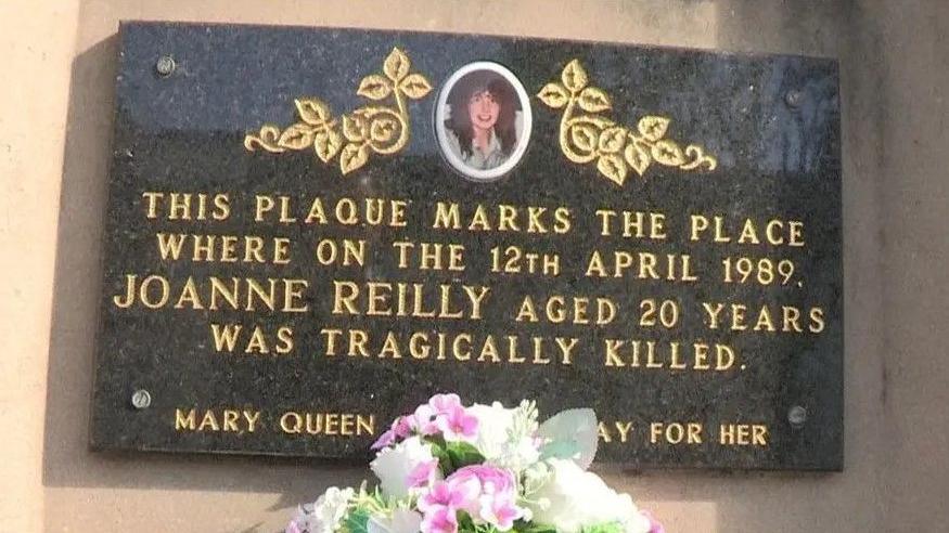 A black plaque with gold writing in memorial of Joanne Reilly. There are white and pink flowers in front of the plaque.