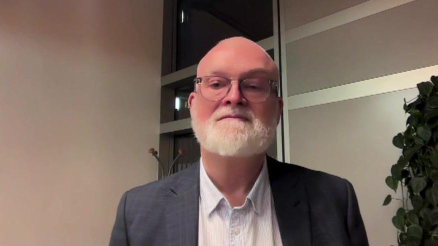 A man with glasses and a beard and wearing a white shirt and grey suit looks into the camera.