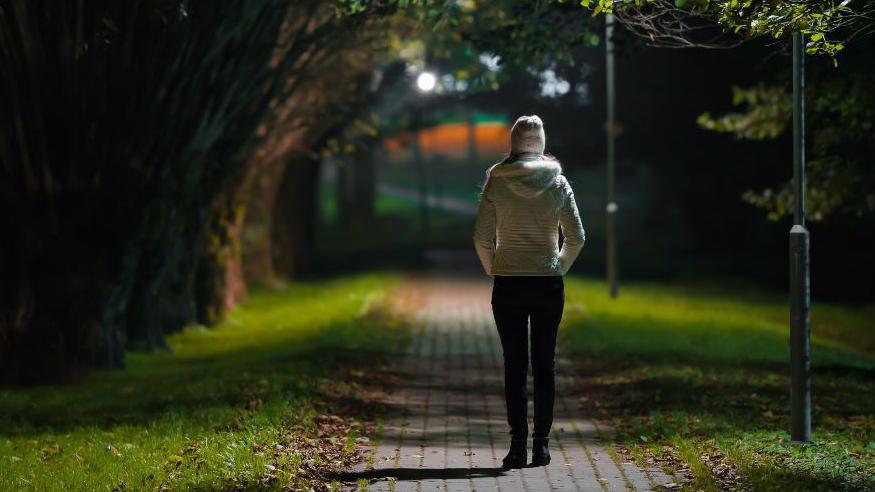 A woman in a white coat and hat walks away from the camera along a dark path between a row of trees and a grassy area.