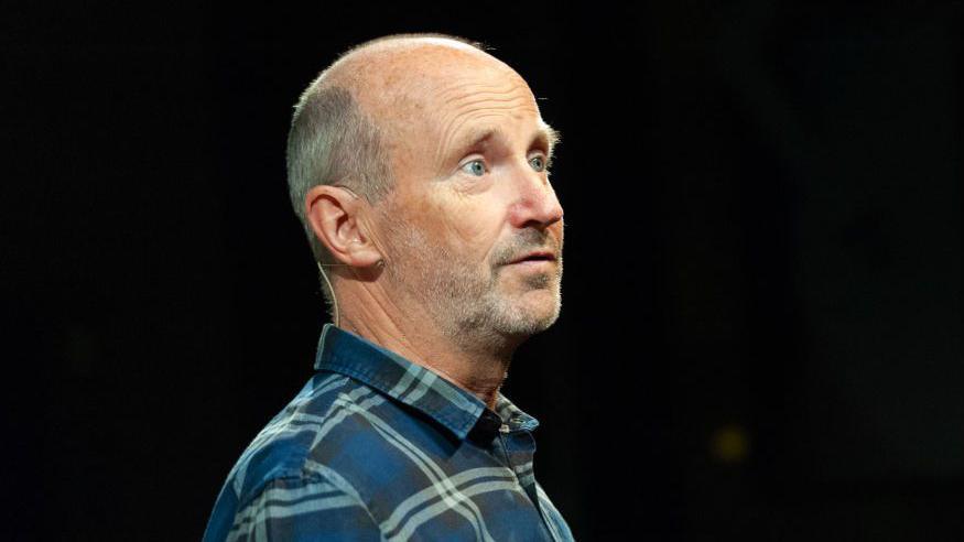 A balding man with short grey hair and a beard is wearing a blue checked shirt