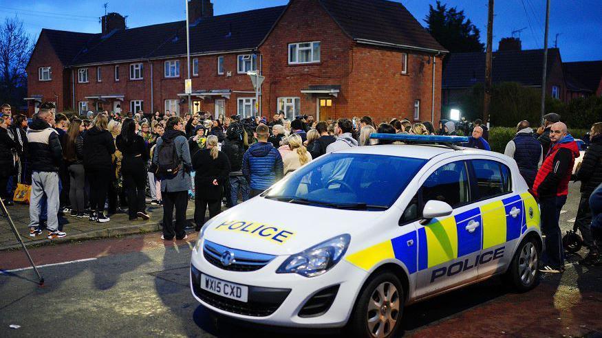 Members of the public observe a minute's silence during a vigil near to the scene in south Bristol where two teenage boys, aged 15 and 16, died after a stabbing attack. A police car is in the foreground. 