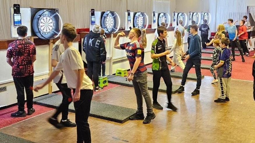 About 20 children of all ages are playing darts. They are in groups of three or four in front of seven darts boards 