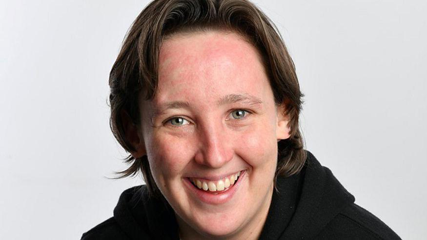 Mhairi Black, a young woman with short loose brown hair, smiles into the camera against a grey backdrop. She is wearing a black hoody.