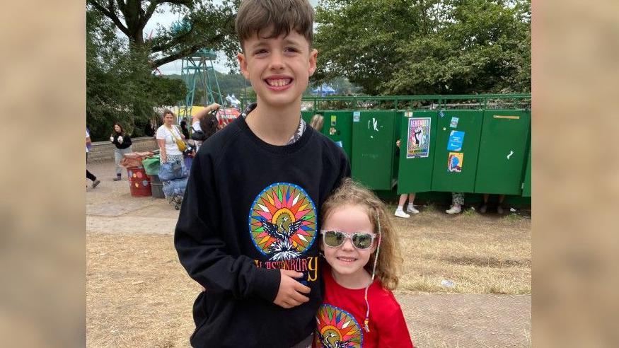 Kelly's children with their arms around each other in front of a bank of porta-loos. Her daughter is in a red Glastonbury top and sunglasses, and her son is in the same top in black.