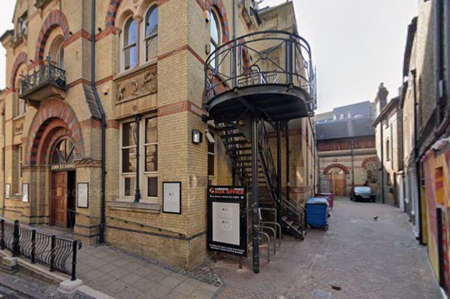 A beige building with "Cambridge Corn Exchange" written above the door. On the right is a passageway with bins, and metal stairs attached to the building