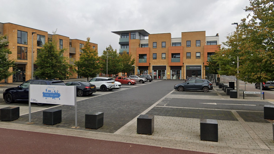 A modern paved square with car parking spaces, cubed bollards, three- and four-story buildings around it, with a sign that reads "THE SQUARE, BROOKLANDS" on the perimeter of the car park.