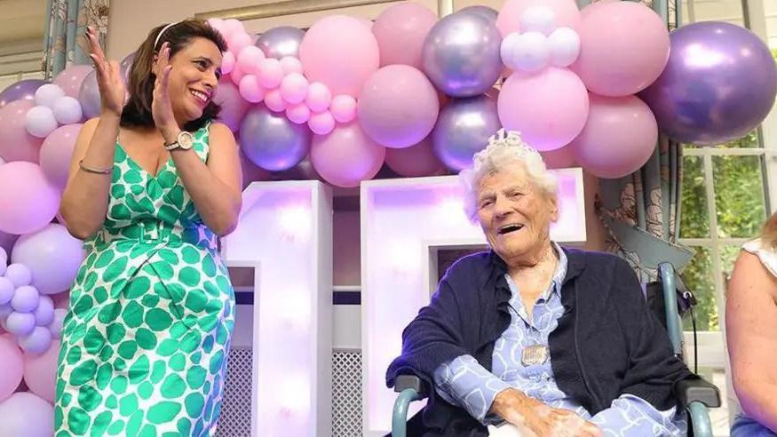 Ethel Caterham sat wearing a tiara with 115 on it with a woman clapping and surrounded by balloons