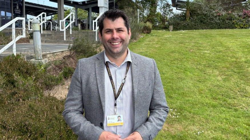 Leigh Frost is smiling as he stands in a garden with County Hall behind him. He has dark hair and brown eyes and is wearing a grey jacket and a grey and white striped shirt and a lanyard with his Cornwall Council identification on it.