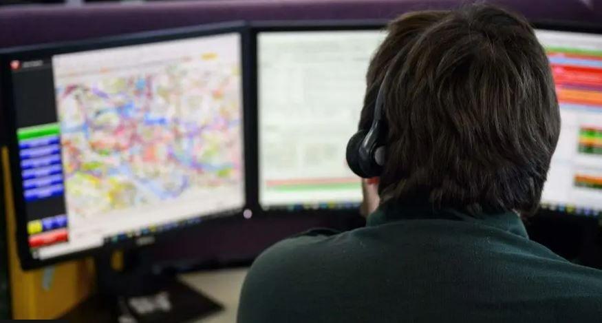 The back of an emergency call handler can be seen. He has brown hair and is wearing a green shirt and a headset. In front of him are computer screens showing digital images that have been blurred out.