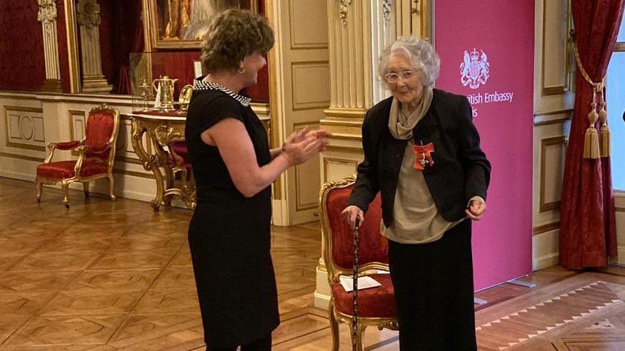 Dame Menna Rawlings stands applauding Noreen Riols in a large, ornate room at the British Embassy in France. Ms Riols, who holds a cane, wears the award on her black jacket. 