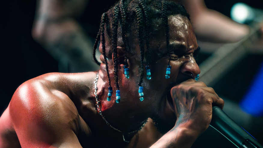 A man with dark coloured braids and blue beads in them sings into a microphone on a stage. He is not wearing a top and is pictured in a crouched position.