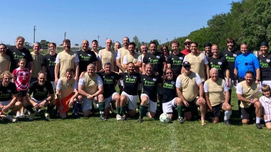 A group of people in the standard football team pre-match photo pose (two rows, with the players in the front row kneeling). Some are wearing black jerseys with "Finn McCool's" emblazoned on them in lime green writing.  Others are wearing gold tops with white sleeves.