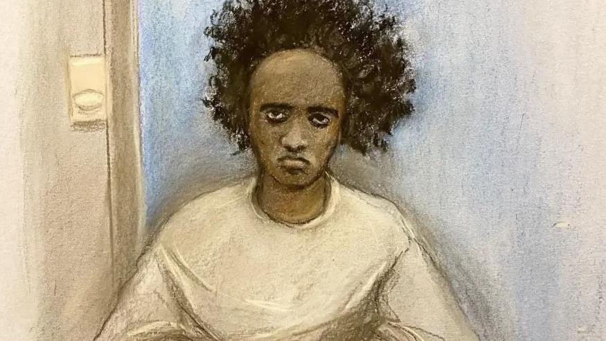 A court sketch of Axel Rudakubana, who has bushy black hair and sits with a blank expression. He is wearing a round-necked white t-shirt.