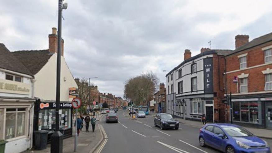 Road with a blue car at the front of the photo, with green traffic lights just beyond