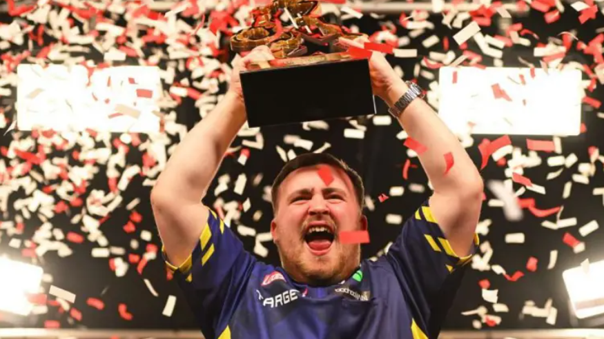 Luke Littler celebrating after his win by holding his trophy aloft, open-mouthed, with a background of red and white confetti falling down around him