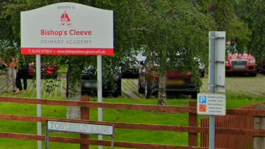 A photo of the sign outside Bishop's Cleeve Primary Academy. The sign is behind a wooden fence and there are some cars parked behind it.