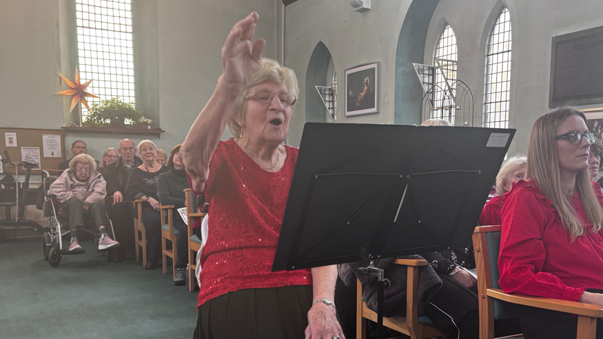 An elderly woman in a red top conducting a choir inside a church