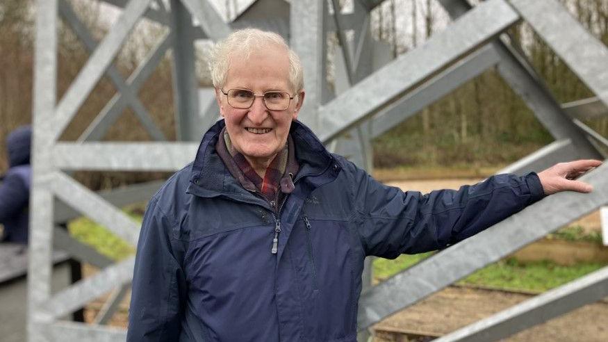 David Anthony Rowley is stood against a metal structure. He has light grey hair and is wearing a navy blue raincoat.