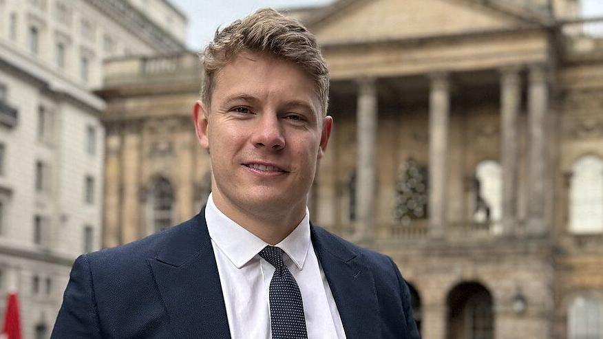 Carl Cashman has short blonde hair and is wearing a navy suit, a white shirt and a navy tie with white dots. He is stood in from of Liverpool's Town Hall.