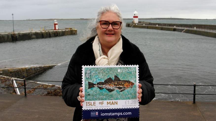 Artist Kimmy McHarrie with a large version of one of her stamps, standing by the sea