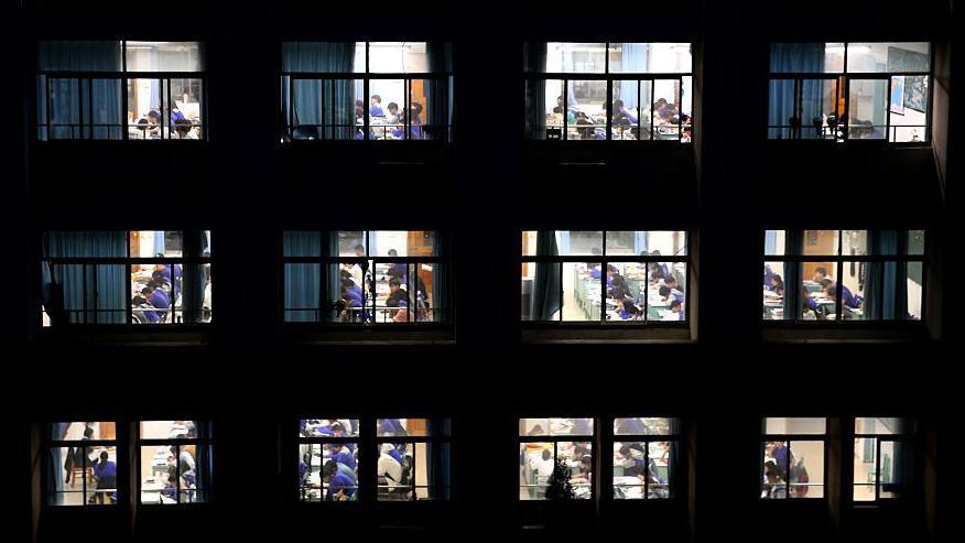 A wide shot of a school building at night, with windows to classrooms showing students studying