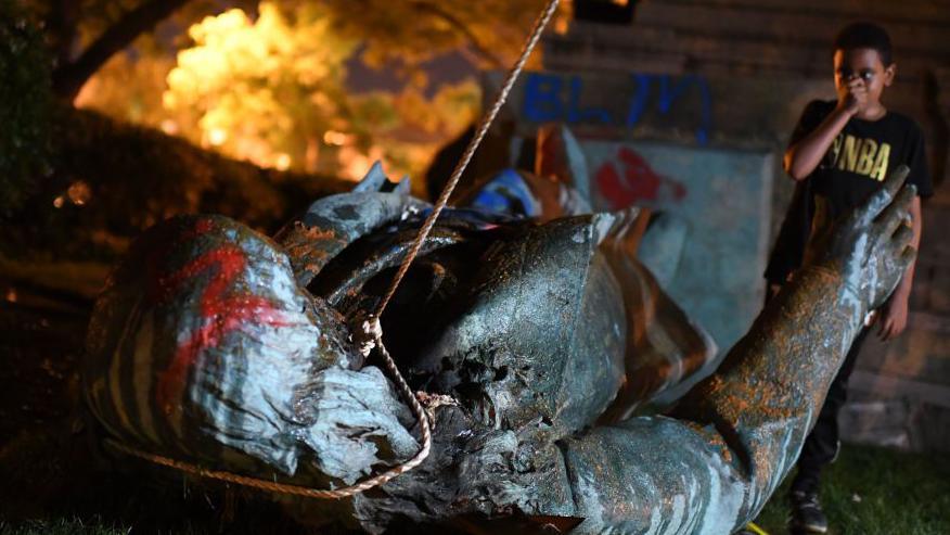 Vandalised statue on the ground as flames blare in the background