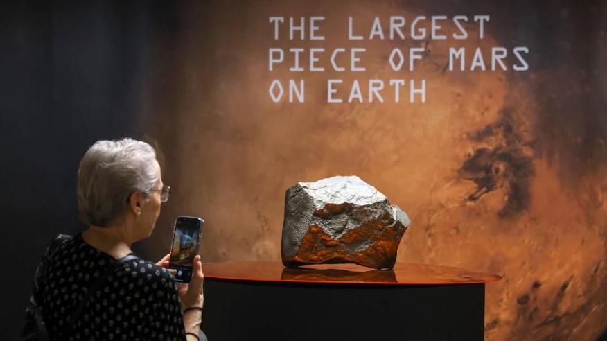 A woman takes a picture of the Martian meteorite with her phone. It glows silver and red and sits under a sign that says "the largest piece of Mars on Earth".
