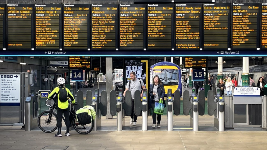 Train station platform gates with cyclist with bike in front looking up at departure board. couple walk through gates at other side with train in background.