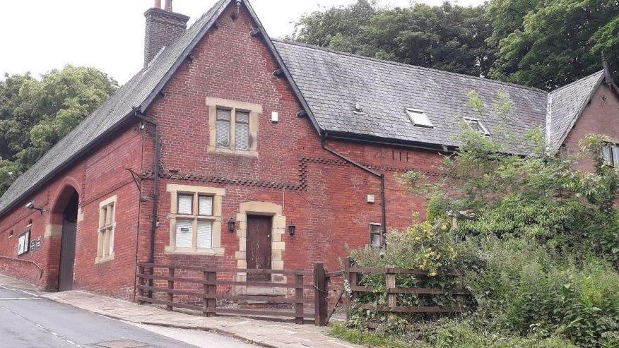 The red-bricked building is situated on the side of a road. There is a wooden fence around the front of the disused heritage centre along with overgrown shrubs and trees.