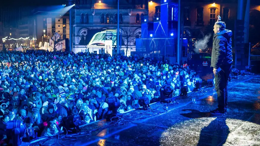 A man stands on on illuminated stage, with a crowd of thousands looking on. There's a blue hue over the whole scene and city centre buildings can be seen in the background.