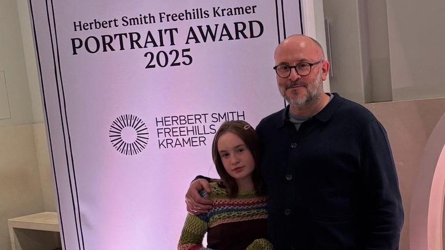Girl with colourful dress and long brown hair stands in front of a banner that reads "Herbert Smith Freehills Kramer Portrait Award 2025". She is stood with a man with glasses and a beard with a dark jumper.