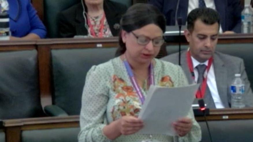 A woman standing in a council chamber, reading from a paper. She is wearing a white dress with green spots and floral embroidery, and has dark brown hair tied back in a centre parting, and glasses. Behind her are rows of green upholstered seats. A man sits in the row behind, in a grey suit, white shirt and grey tie, with a water bottle in front of him.