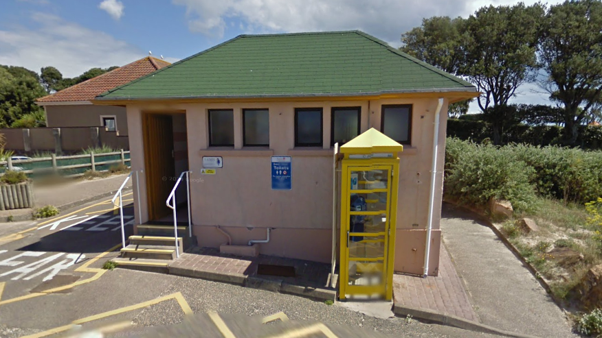 A mid shot of the Green Island public toilets. It is a small hut with a green roof, cream coloured walls, and a yellow phone box outside.
