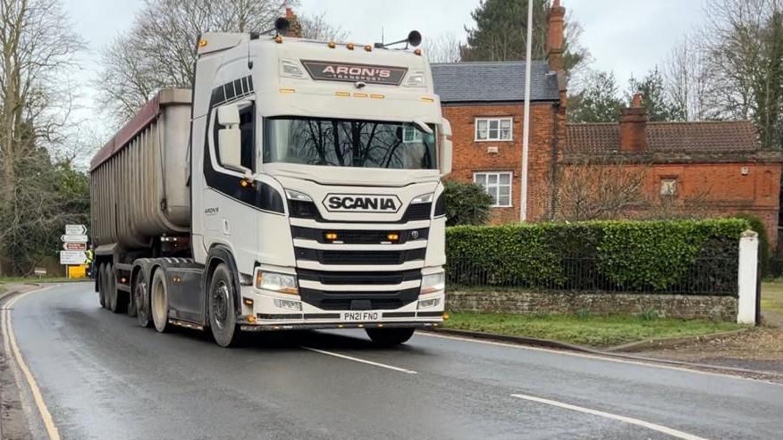 A white Scania lorry driving through Coltishall on the B1150. In the backdrop to the right is a house, and to the left, at the rear of the lorry, is a junction to Great Hautbois (which is pronounced Hobbis).