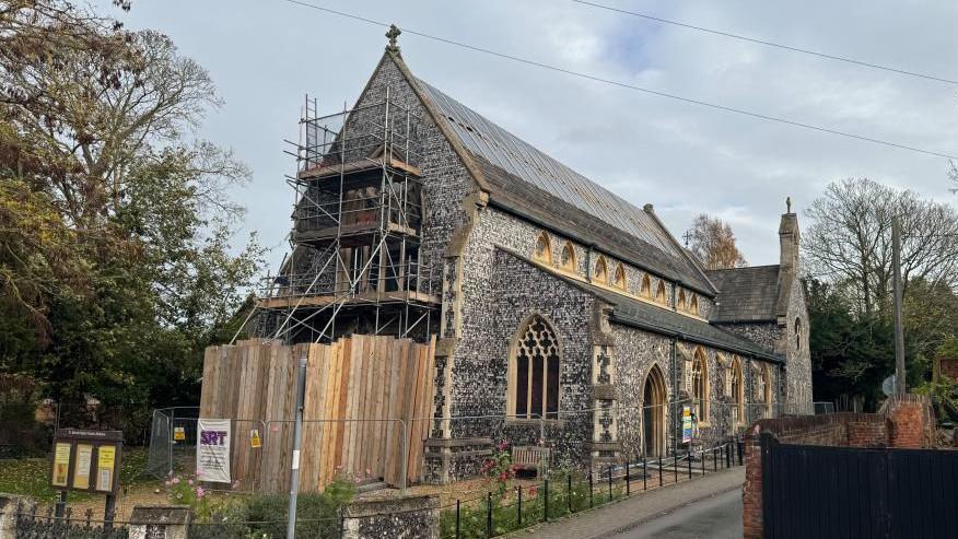 A Victorian church built of square knapped flint, with arched windows with sandstone mullions and a slate roof. The building has been damaged by fire, with plastic sheeting and wooden battens holding temporary repairs in place on the roof. Fencing can be seen around the building, with trees behind.