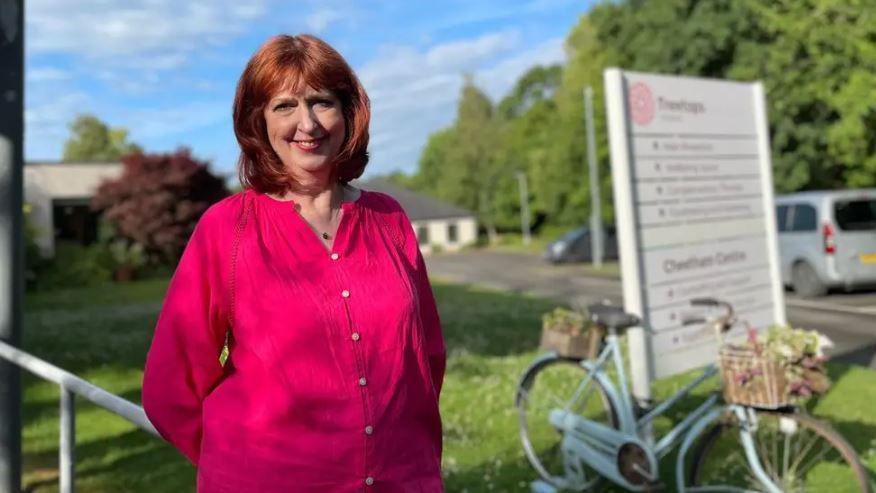 Picture of Julie Heath. She is wearing a dark pink shirt in front of a Treetops Hospice sign.