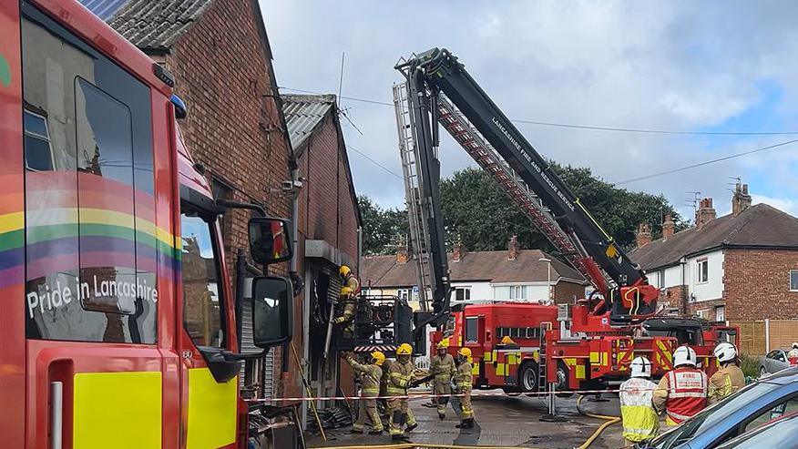 Firefighters inside a cordon tackling a fire at a storage unit on an industrial estate with two fire engines one with an aerial platform close by and a row of houses nearby.