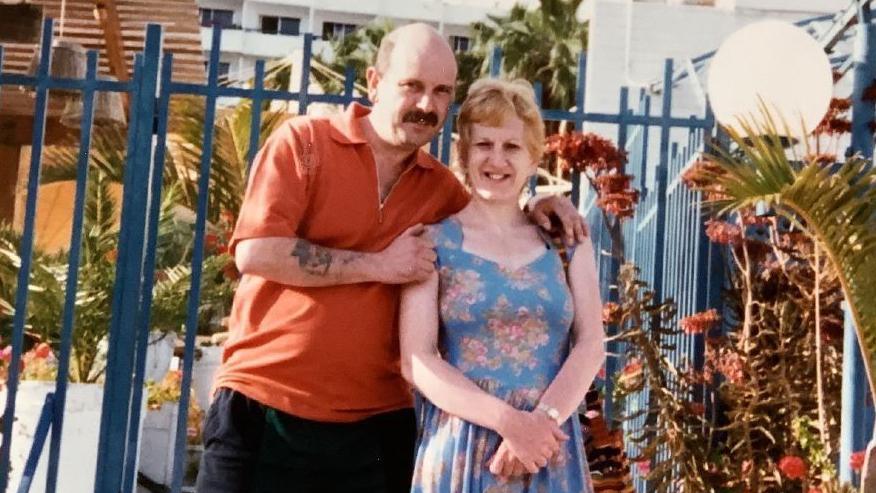 Tony Hutchinson and Wendy Hutchinson pictured on holiday. Wendy is wearing a blue dress with floral patterns, with Tony in an orange top and dark trousers. They are stood by blue iron gates and are surrounded by exotic plants. 