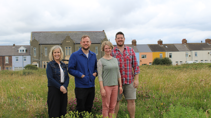 Four people stand in the wild grass in front of a row of coloured houses which make up the main street of the coastal village of Cambois.