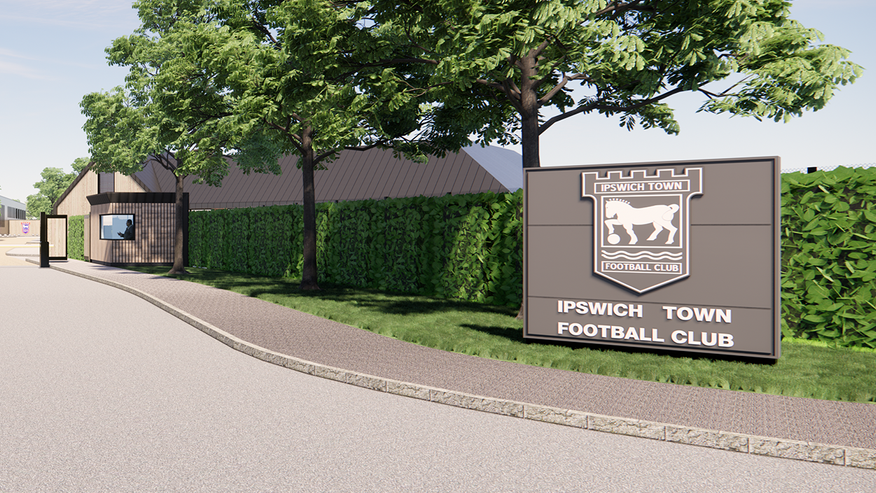 Roadway onto site, with pavement and large "Ipswich Town Football Club" sign. There are trees and a hedge lining the route into the training ground. A security gatehouse is visible by the gate to the left of the picture.