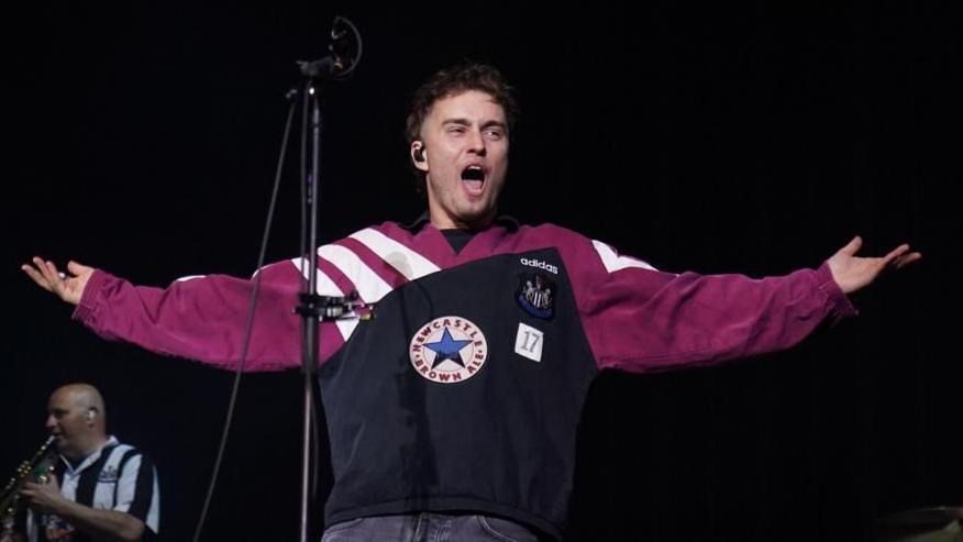 The singer Sam Fender stands with his arms open singing. He is wearing a purple NUFC top with the words Newcastle Brown Ale on it.