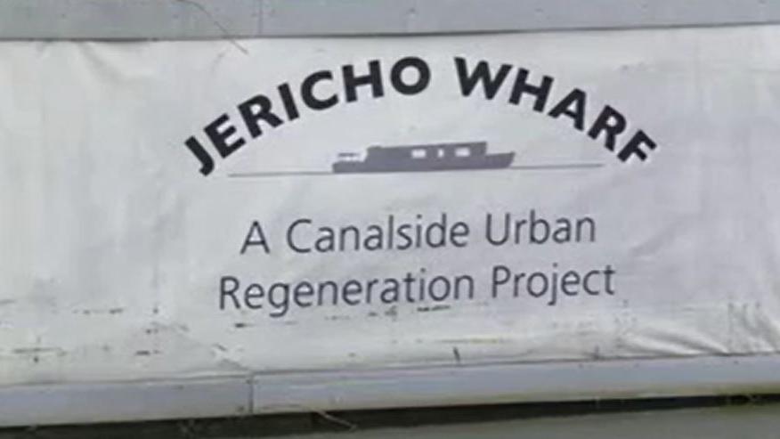 A white placard reading "Jericho Wharf, A Canalside Urban Regeneration Project". There is a drawing of a boat in between the writing.