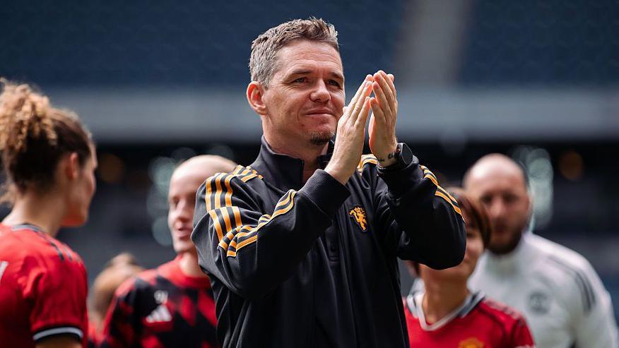 Marc Skinner, in black tracksuit top with orange stripes, applauds with players and staff in the background