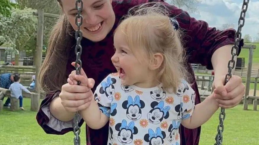 Chelsea Gleason-Mitchell smiles at her daughter as she stands behind her and pushes her on a swing. Isabella is laughing and wearing a T-shirt with Minnie Mouse on it. 