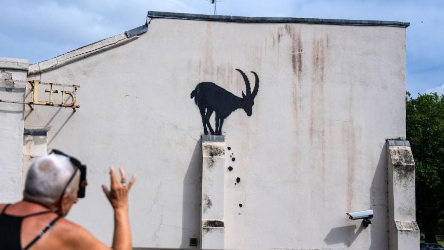 The Goat artwork is seen on the side of a white washed building. It depicts a silhouette of a large-horned goat perched on top of a buttress with painted rocks coming down. A woman is seen in the corner of the image taking a picture.