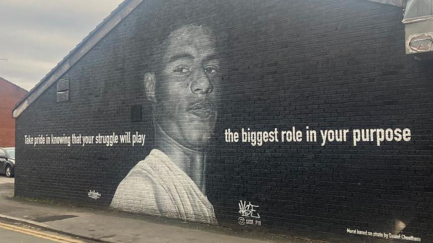 The mural of Marcus Rashford in Withington, Manchester, painted against the side of a building. It portrays the footballer, in a white t-shirt, alongside the words: 'Take pride in knowing that your struggle will play the biggest role in your purpose'. 