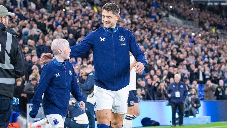 Mason, who is wearing an Everton FC kit, is walking on to the pitch alongside James Tarkowski. Crowds of spectators can be seen in the background. 